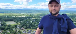 Iskandar Sattibaev smiling at the camera with green hills and forrest in the background