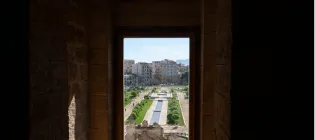 Window overlooking a square with trees lined up in rows on a sunny day. In the background, there are apartment complexes, cars parked and a mountain in the distance. 