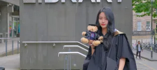 Student Chengcheng in graduation robes in front of the library, holding a teddy bear
