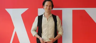 Photo of Patrick Errington holding his Emerging Writers award in front of a red and white screen