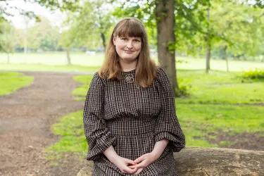 Photo of Suzanne sitting in a parkland setting. She is smiling at the camera.