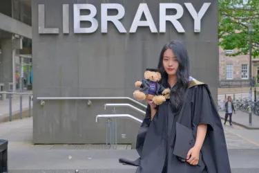 Student Chengcheng in graduation robes in front of the library, holding a teddy bear