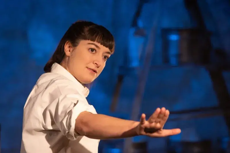 a woman on stage in a white shirt stares off into the distance with her hand raised in a steadying motion