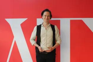 Photo of Patrick Errington holding his Emerging Writers award in front of a red and white screen