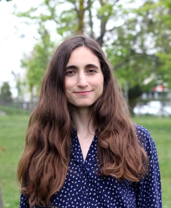 Photo of Dr Hamide Elif Üzümcü, who is smiling. She is standing in a park with trees behind her.